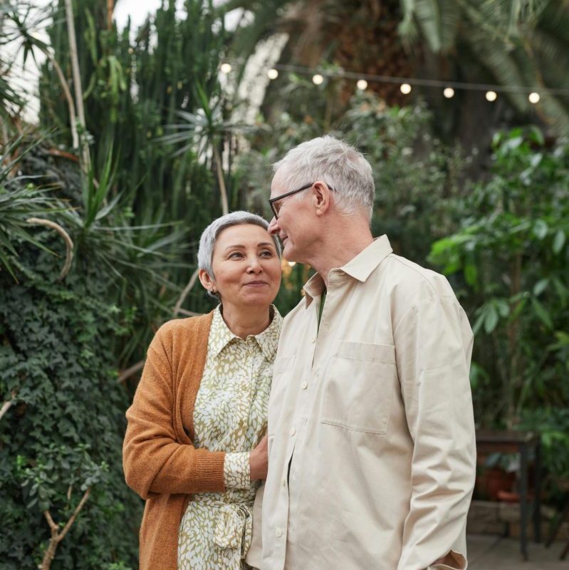 A senior couple shares a tender moment in a vibrant greenhouse filled with lush plants.